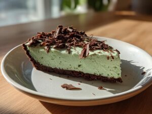 A close-up of a slice of light green grasshopper pie with a dark chocolate crust, topped with chocolate shavings.
