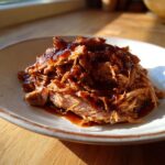 A close-up of tender, shredded Slow Cooker Pulled Pork piled high and generously drizzled with dark BBQ sauce on a speckled plate.