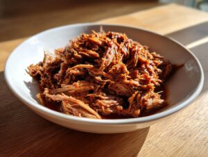 A white bowl filled with shredded, saucy Slow Cooker Pulled Pork, highlighted by sunlight.