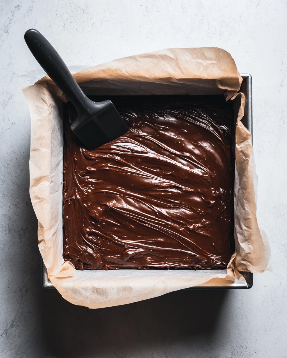 Overhead view of smooth, melted chocolate mixture being spread in a square pan for making fudge.