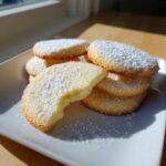 A stack of soft sugar cookie rounds dusted heavily with powdered sugar, one cookie has a bite taken out showing the soft interior.