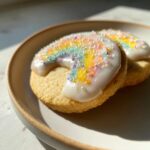Close-up of two soft sugar cookies topped with white icing shaped like a rainbow and colorful sprinkles.
