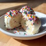 A close-up of two halves of a sugar cookie truffle showing the dense interior, coated in white chocolate and rainbow sprinkles.