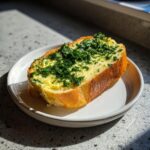 A thick slice of toasted bread generously topped with melted garlic butter and fresh green herbs.
