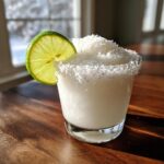 A frosty white christmas margarita topped with shredded coconut and garnished with a lime wheel.