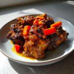 Close-up of rich, dark brown stew chicken pieces topped with bright red pepper chunks on a light blue plate.
