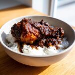 A close-up of a piece of rich brown stew chicken served over fluffy white rice in a light-colored bowl.