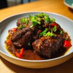 Close-up of rich, dark brown stew chicken Jamaican recipe pieces in a savory sauce, topped with green onions.