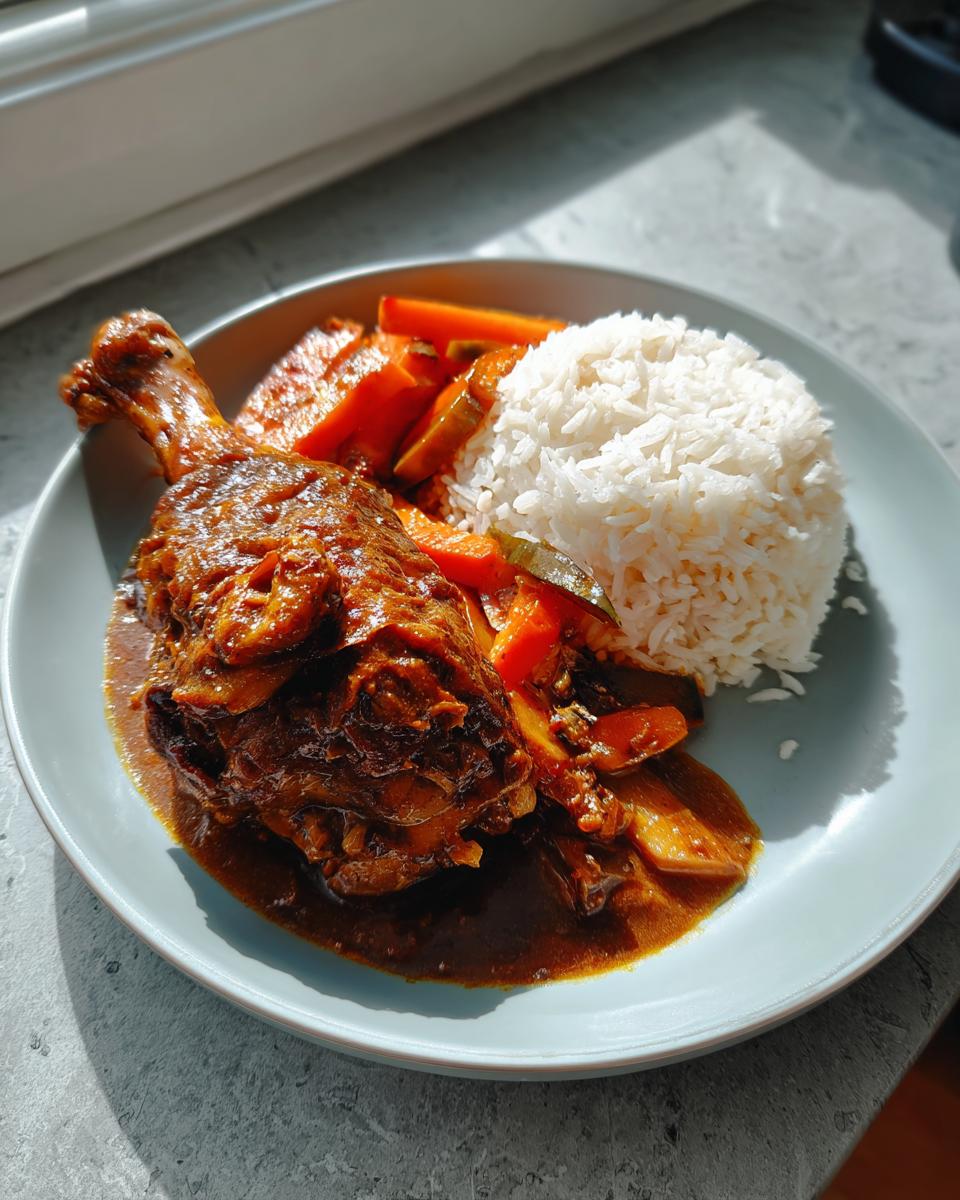 A plate featuring a serving of rich brown stew chicken leg quarter next to white rice and cooked carrots.