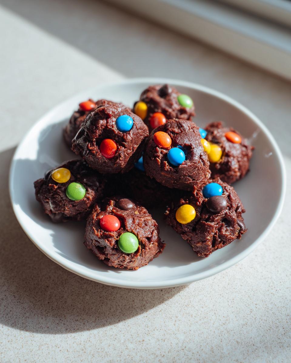 A white plate holding several rich, dark chocolate cosmic brownie protein bites topped with colorful candy pieces.