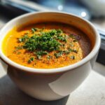 A close-up of a bowl of creamy, orange roasted vegetable soup topped with fresh chopped parsley.