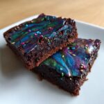 Two rich, fudgy gluten free cosmic brownies stacked slightly, featuring vibrant, marbled galaxy icing and colorful sprinkles.