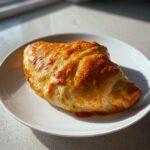 A single, golden-brown baked cheeseburger pocket resting on a white plate, showing flaky pastry.
