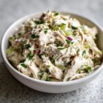 Close-up of shredded chicken salad mixed with celery, red onion, and herbs in a white bowl.