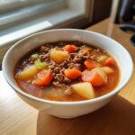 A close-up of a steaming bowl of hamburger vegetable soup filled with ground beef, carrots, and potatoes.
