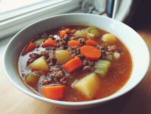 Close-up of a white bowl filled with rich hamburger vegetable soup, featuring ground beef, carrots, potatoes, and celery.