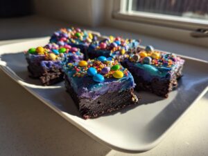Four squares of rich, dark healthy cosmic brownies topped with vibrant blue/purple frosting and candy pieces.