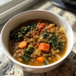 Close-up of a white bowl filled with hearty lentil vegetable soup featuring carrots, kale, and tomatoes.