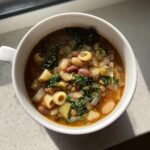 Overhead view of a bowl filled with hearty vegetable soup containing beans, lentils, kale, and small pasta shapes.