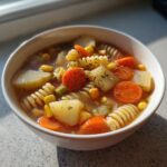 Close-up of a bowl of hearty vegetable soup featuring carrots, potatoes, corn, and rotini pasta.