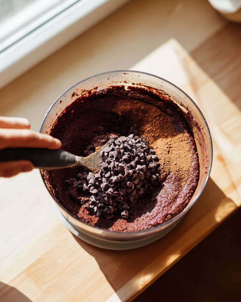 A hand adds mini chocolate chips and cocoa powder to a thick batter for healthy cosmic brownies.