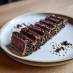 Slices of rare, sesame-crusted pan seared tuna steak arranged on a light plate with scattered sesame seeds.