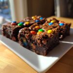 Four squares of fudgy protein cosmic brownies topped with colorful candy-coated chocolates, served on a white plate.