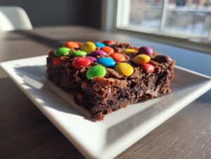 A rich, fudgy square of protein cosmic brownies topped with colorful candy-coated chocolates, sitting on a white plate.