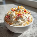 A close-up of a fluffy cream cheese dip for crackers, topped with shredded cheddar, red peppers, and chives.