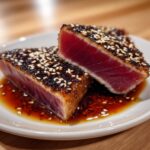 Two thick slices of perfectly seared, sesame-crusted grilled tuna steak showing a rare, deep red center, resting in soy sauce.