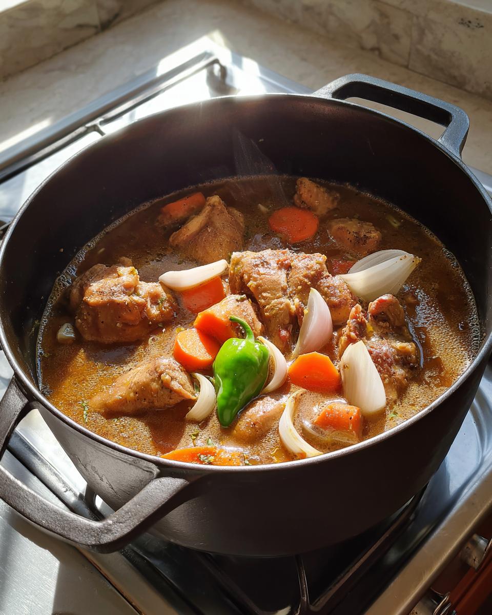 Close-up of brown stew chicken simmering with carrots, onions, and a whole green pepper in a black pot.