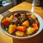 A close-up of a hearty bowl of vegetable soup with stew meat, potatoes, carrots, and peas in a rich brown broth.